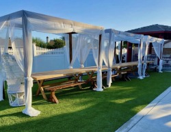 Outdoor Los Angeles graduation party rentals setup featuring wooden farm tables, benches, and an elegant white draped canopy tent on a lawn